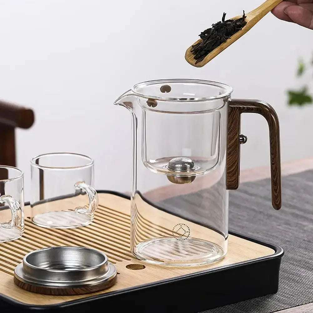 Théière en verre avec poignée en bois, dotée d’un infuseur en inox. Une cuillère en bois verse des feuilles de thé dans la théière, posée sur un plateau en bois avec des tasses assorties.