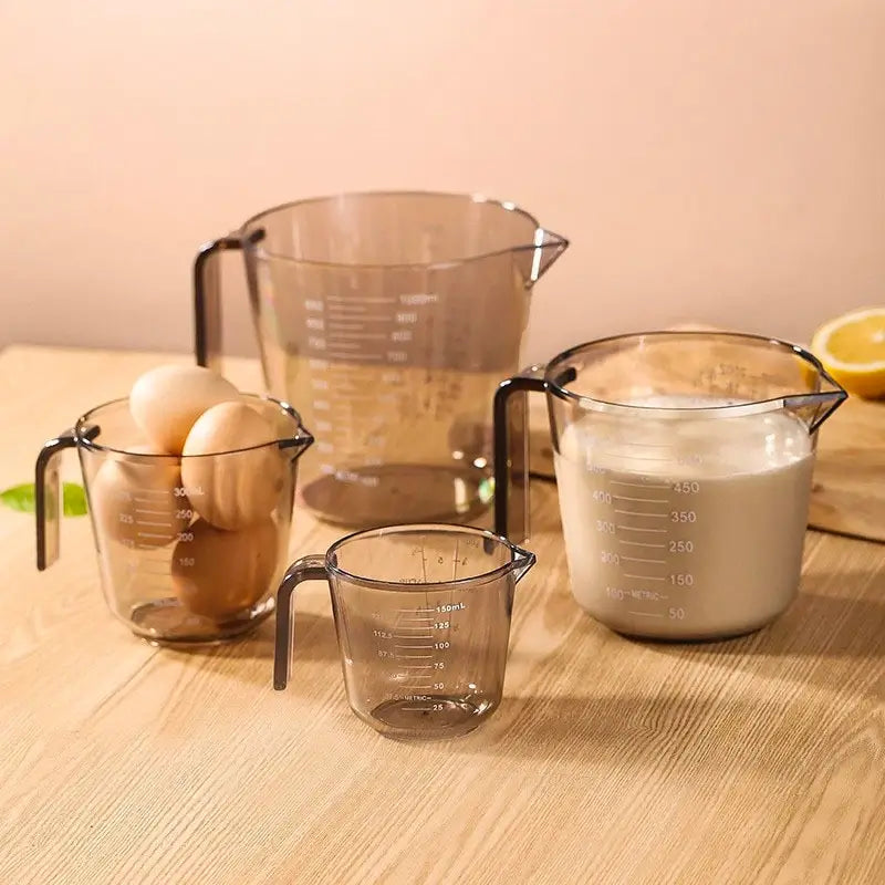 Un ensemble de tasses à mesurer transparentes de différentes tailles (150 ml, 300 ml, 500 ml et 1 litre), contenant des œufs et du lait, disposées sur une table en bois.