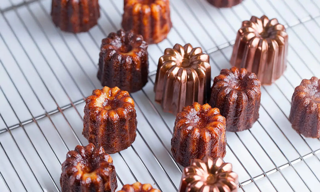 Cannelés dorés disposés sur une grille de refroidissement avec des moules en cuivre visibles parmi les pâtisseries.