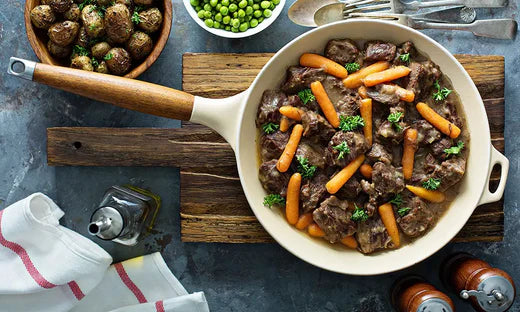 Plat de pot au feu servi dans une poêle blanche, garni de morceaux de bœuf, de petites carottes et de persil, accompagné de pommes de terre rôties et de petits pois en arrière-plan.