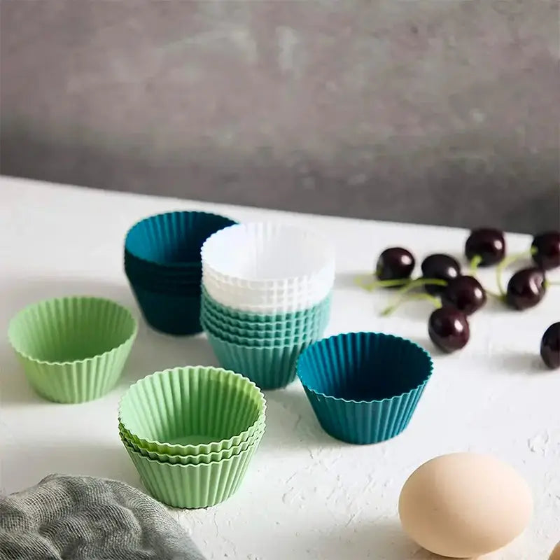 Une disposition artistique de moules à cupcakes en silicone sur un plan de travail blanc avec des cerises et un œuf en arrière-plan.