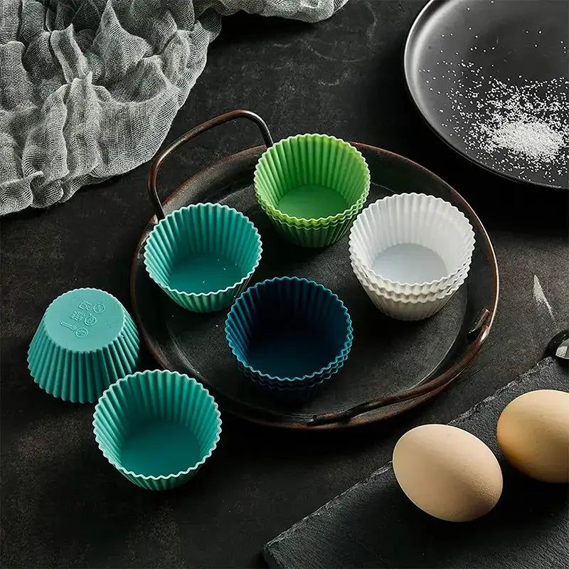 Des moules à cupcakes en silicone de couleurs variées (bleu, vert, blanc) disposés sur une assiette sombre, avec des accessoires de cuisine comme des œufs en arrière-plan.