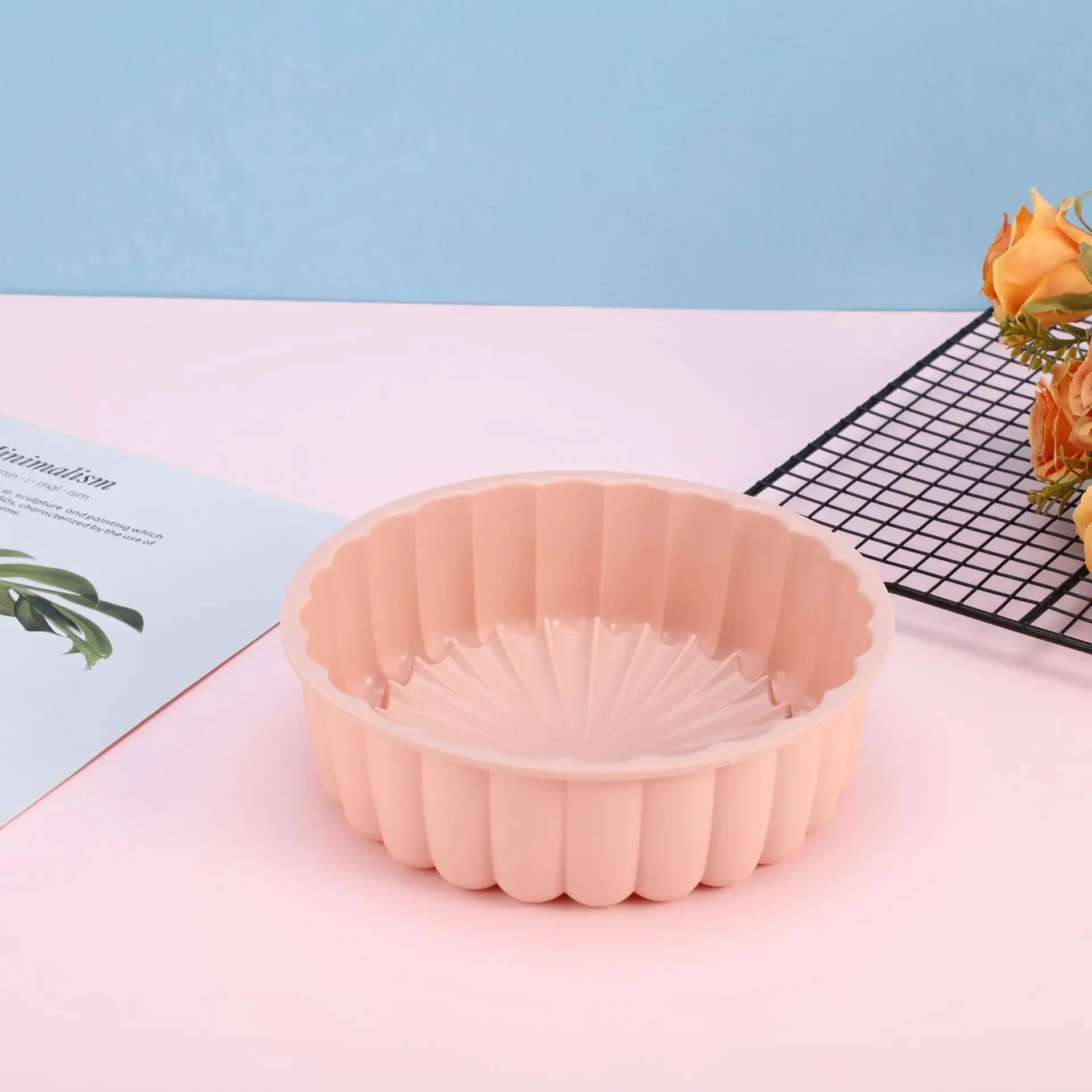 Moule à gâteau en silicone rose clair, de forme ronde avec des bords ondulés, posé sur une surface rose avec une grille noire et un bouquet de fleurs en arrière-plan.