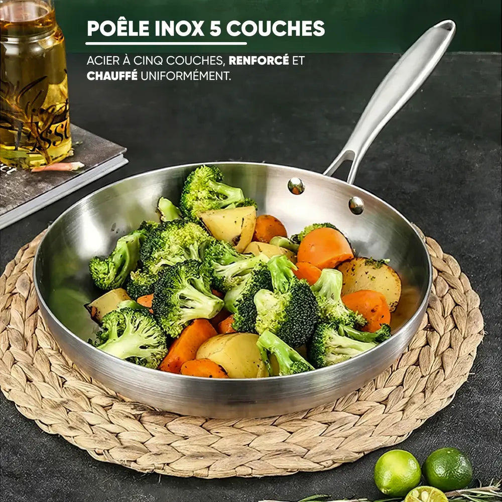 Une poêle en acier inoxydable à 5 couches contenant des légumes sautés, dont des brocolis, des carottes et des pommes de terre, placée sur un dessous de plat en osier.