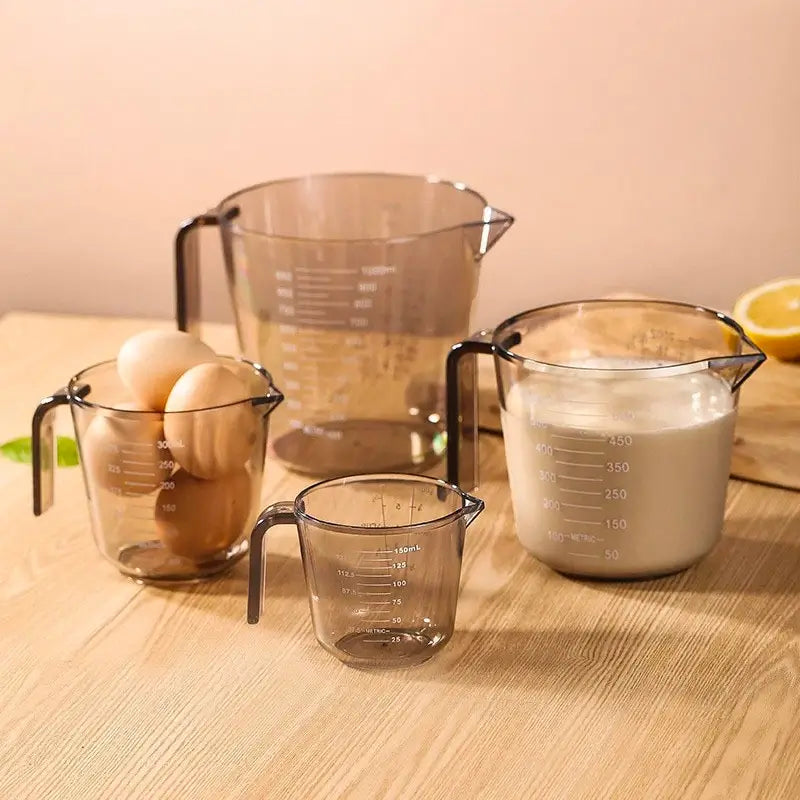 Un ensemble de tasses à mesurer transparentes de différentes tailles (150 ml, 300 ml, 500 ml et 1 litre), contenant des œufs et du lait, disposées sur une table en bois.