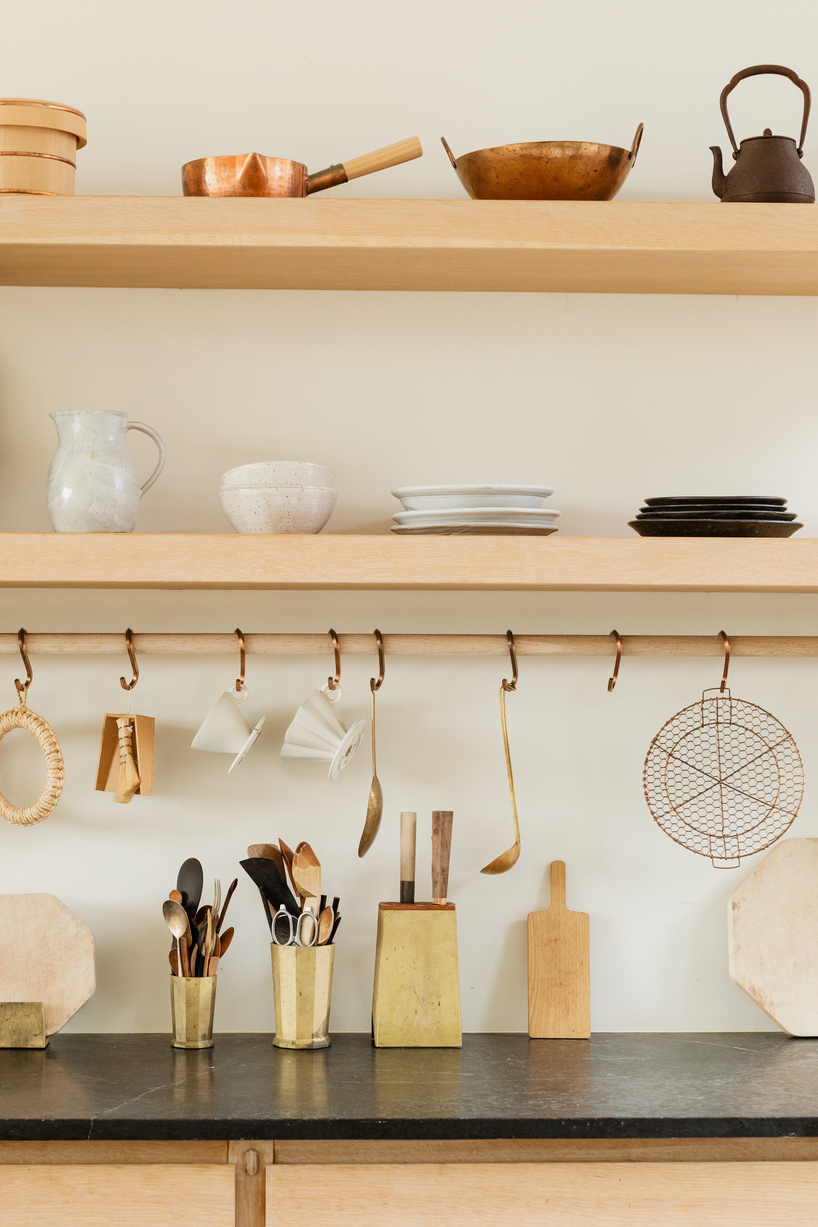 ne cuisine élégante avec des étagères en bois clair contenant divers ustensiles de cuisine en cuivre et en céramique. Sur le plan de travail, il y a un bloc de couteaux en bois, des pots contenant des ustensiles de cuisine, et d’autres accessoires de cuisine. Les ustensiles sont suspendus à une barre en métal sous l’étagère inférieure. Des assiettes et des plats sont soigneusement empilés sur les étagères supérieures.