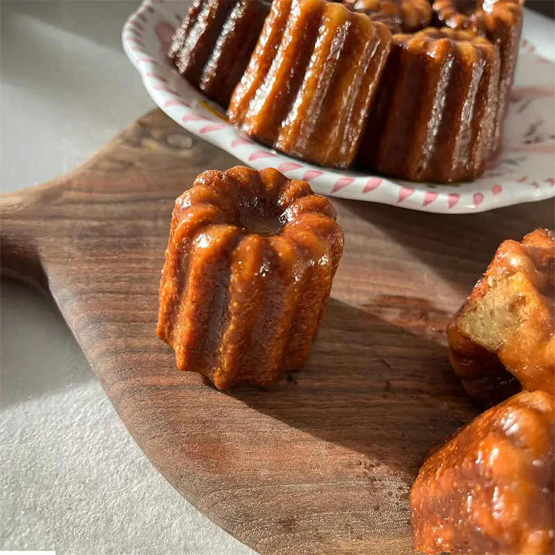 Zoom sur un cannelé maison doré et caramélisé, posé sur une planche en bois avec d'autres cannelés visibles en arrière-plan.