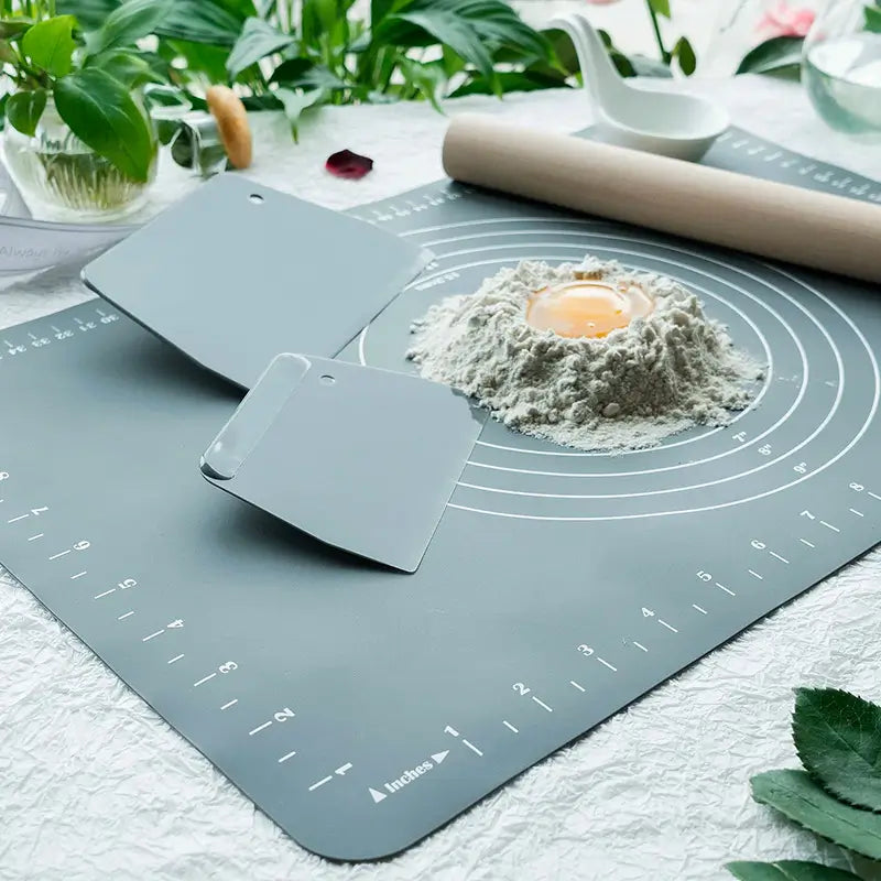 Un tapis de pâtisserie en silicone gradué avec deux cornes de pâtissier grises, un monticule de farine contenant un jaune d'œuf, et un rouleau à pâtisserie en bois en arrière-plan, entouré de plantes vertes.