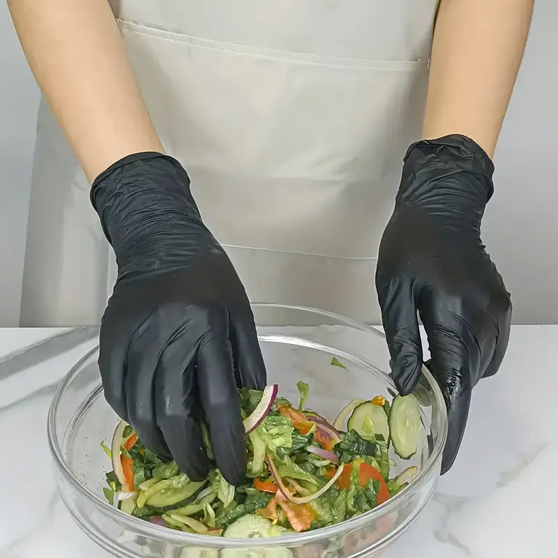 Une personne portant des gants noirs mélange une salade de légumes frais dans un bol en verre, portant un tablier beige."