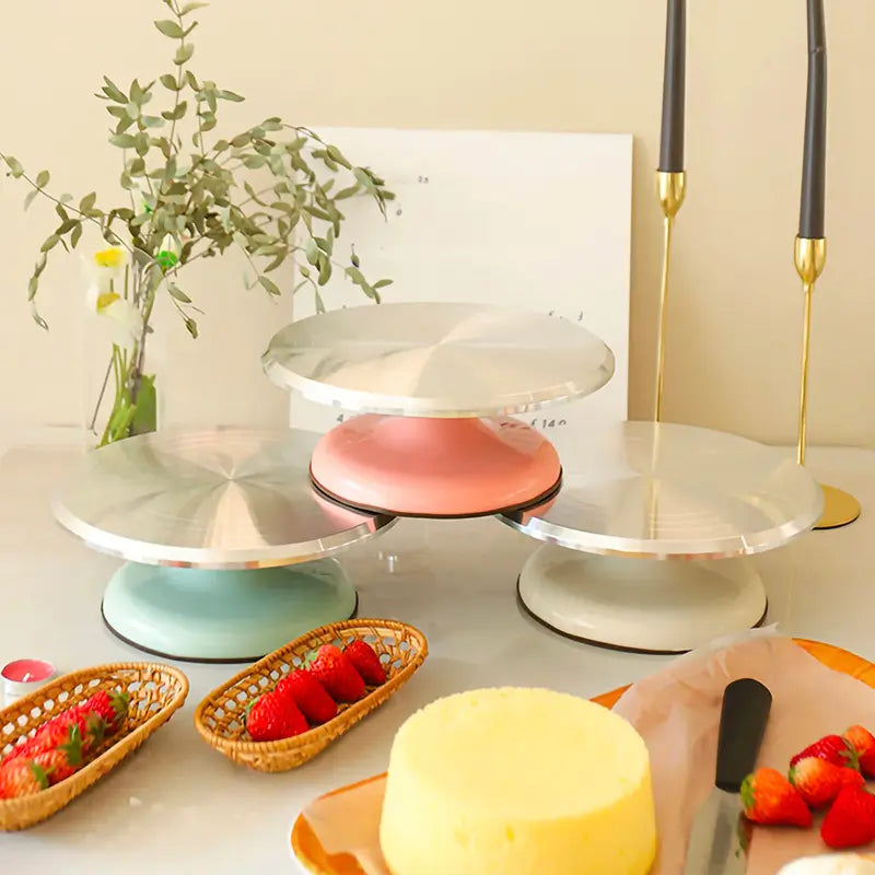 Trois plateaux tournants en métal avec des bases colorées (rose, vert et blanc) disposés sur une table avec des fraises fraîches, un gâteau jaune et des éléments de décoration comme des bougies et des plantes.