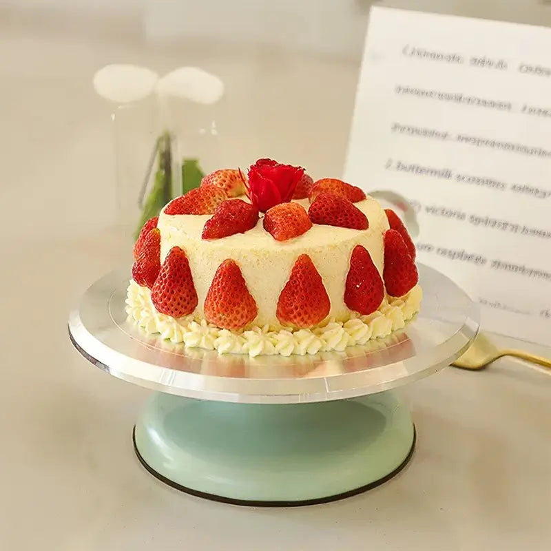 Un gâteau rond recouvert de crème et décoré de fraises entières autour et sur le dessus, avec une rose rouge au centre, présenté sur un support de gâteau en métal et céramique.