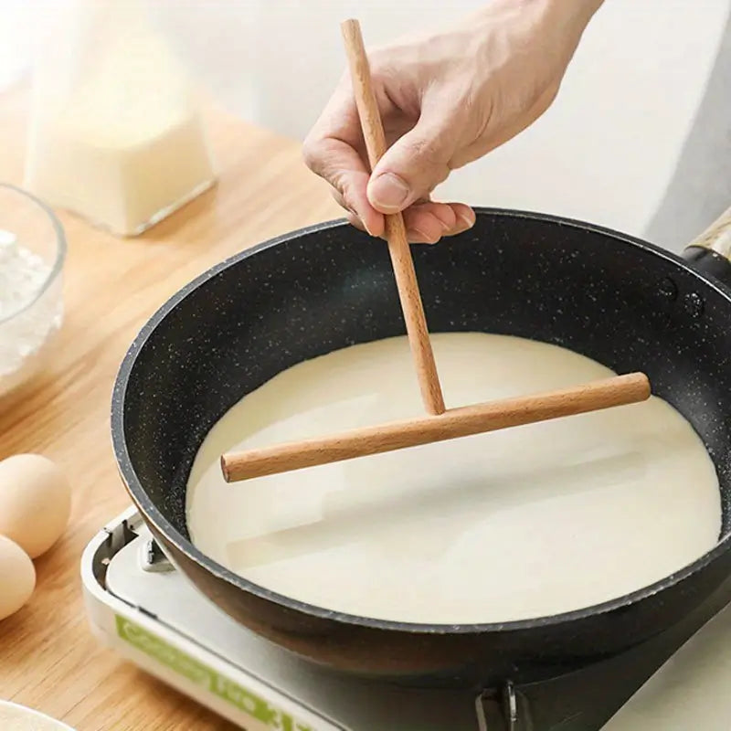 Une main utilisant un râteau à crêpes en bois pour étaler de la pâte dans une poêle noire, illustrant son usage pratique.