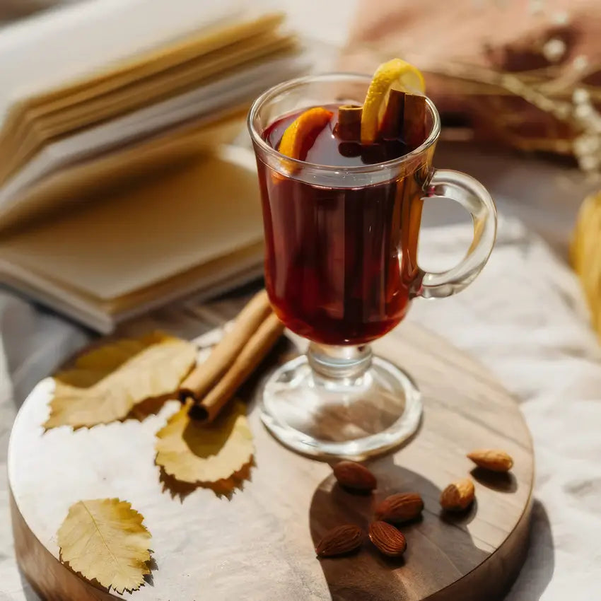 Verre de Irish coffee ou boisson chaude épicée avec des tranches d'orange et un bâton de cannelle, posé sur une planche en bois, entouré de feuilles sèches et d'amandes, avec un livre ouvert en arrière-plan